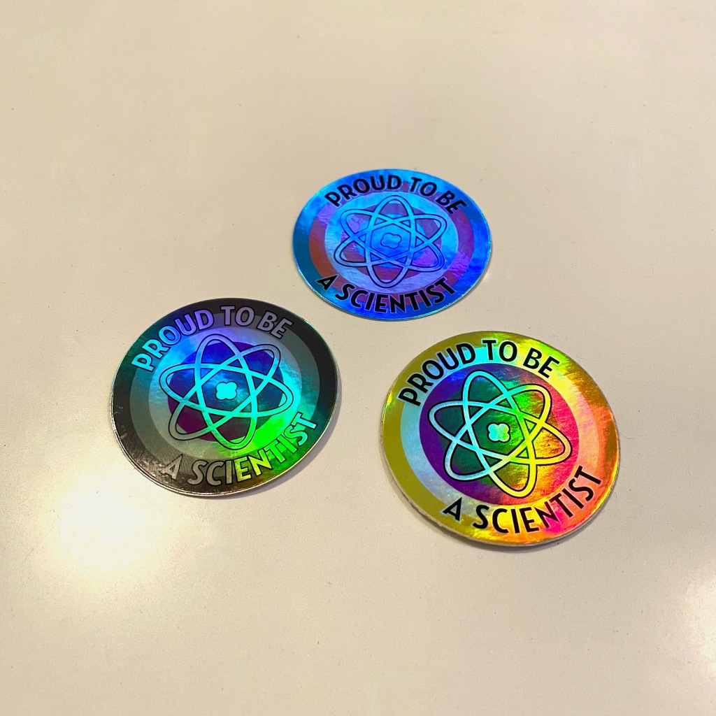 A photo of three circular stickers on a cream-coloured surface. Each sticker has "Proud to be a scientist" written on it in a ring around a 3-oval atom symbol, and are coloured according to the trans, non-binary, and asexual flag colours. All the stickers are shiny and are reflecting different colours of light depending on angle.